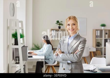 Ritratto di una donna d'affari bionda sorridente e sicura in tuta grigia in piedi con le braccia piegate in ufficio. Foto Stock