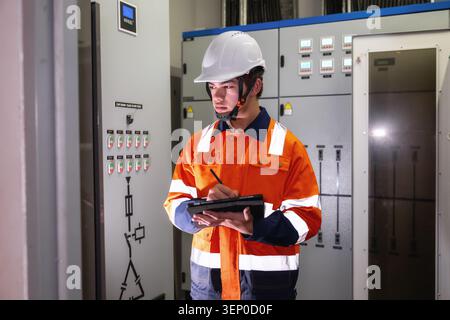 Giovane ingegnere industriale asiatico in gilet di sicurezza arancione e supporti per elmetto bianco. contiene tablet e penna. esamina lo schermo con attenzione. Pannelli elettrici lin Foto Stock