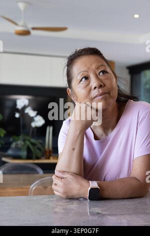 Donna asiatica anziana seduta al bancone in pietra della cucina, che riposa il mento, indossa T-shirt rosa e smartwatch Foto Stock