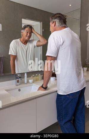 Uomo maturo in piedi al lavandino del bagno che regola i capelli grigi indossando T bianca mentre si guarda nello specchio Foto Stock