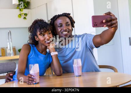 Coppie diverse che si appoggiano al tavolo della cucina di casa, scattano selfie con lo smartphone rosa e i frullati Foto Stock