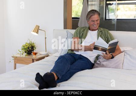 Uomo maturo sdraiato sul letto indossando un orologio da polso che legge un libro cartaceo aperto a casa con tè Foto Stock