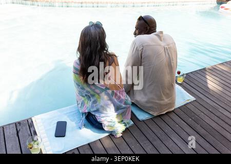 Coppie diverse sedute su asciugamani a bordo piscina su una terrazza in legno, smartphone e cocktail nelle vicinanze Foto Stock