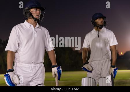 Diversi giocatori di cricket maschi che indossano divise bianche in piedi sull'erba al crepuscolo, tenendo pipistrelli, caschi, piazzole Foto Stock