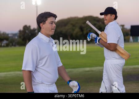 Due giocatori di cricket maschi adulti in piedi sul campo di cricket al crepuscolo indossando una divisa bianca con pipistrelli e guanti Foto Stock