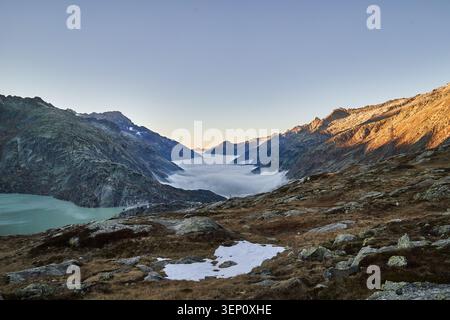 La luce dell'alba bagna aspre vette alpine e un lago turchese, mentre la bassa nebbia riempie l'ampia valle. Foto Stock