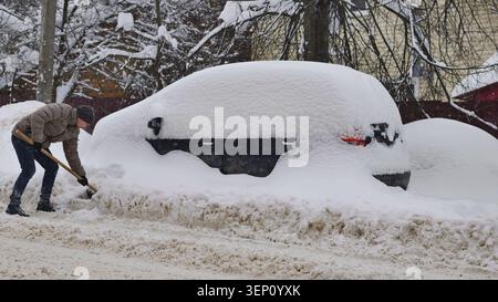 Uomo che spara neve dalla sua auto sepolta in una strada dopo una dura tempesta invernale Foto Stock