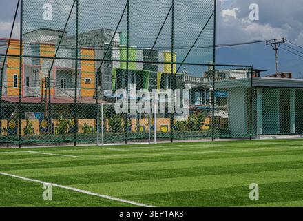 Palu, Indonesia - 22 maggio 2026: Un campo di calcio verde circondato da una recinzione a filo alto. I gol bianchi sono pronti per giocare sotto un cielo nuvoloso. Edifici colorati incorniciano lo sfondo, aggiungendo un contrasto urbano. Questo campo da calcio in miniatura è situato nel centro della città. Foto Stock