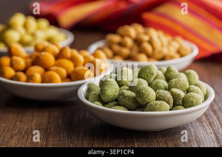 Arachidi rivestite assortite servite in piccole ciotole bianche su un tavolo di legno, caratterizzate da croccanti e aromatizzati in giallo, arancione e verde. Ciotole colorate ideali per feste, antipasti o spuntini informali. Foto Stock