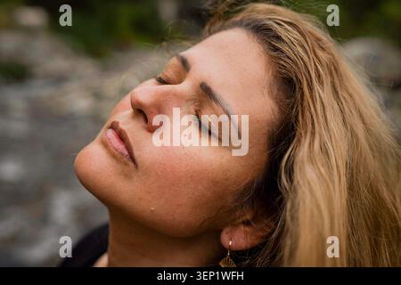 Donna bionda che chiude gli occhi e inclina la testa verso l'alto, sperimentando un momento di consapevolezza e serenità mentre medita all'aperto in una calma naturale Foto Stock