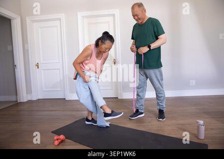 Coppia asiatica che indossa abbigliamento sportivo a casa, tiene il metro a nastro, donna anziana che tira pantaloni sul tappetino Foto Stock