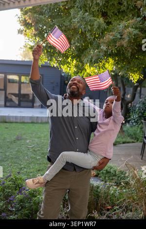 Padre afroamericano maturo che tiene in mano un bambino e sventola bandiere americane nel cortile con una camicia blu Foto Stock