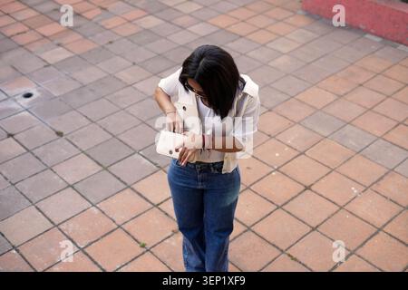 Giovane donna in piedi all'aperto su piastrelle di terracotta quadrate, vestita in modo informale con jeans e una camicia bianca, guardando meticolosamente attraverso il suo piccolo Foto Stock