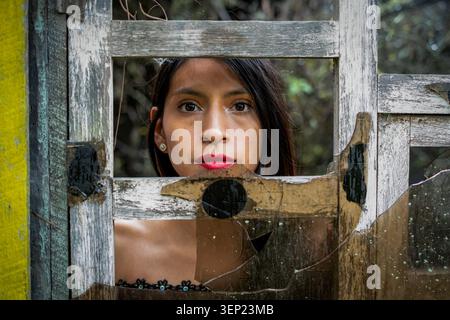 Giovane donna con labbra rosse che sbircia attraverso una vecchia cornice di legno della finestra con vetro rotto, esprimendo sentimenti di tristezza e contemplazione mentre si connette Foto Stock