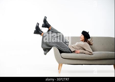 Donna sdraiata sul divano con gambe rialzate e sorridente in studio. Giovane donna caucasica che indossa berretto nero, maglione beige, pantaloni larghi grigi, stivali neri, umore rilassato e giocoso Foto Stock