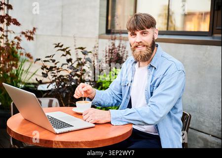 Ritratto di un professionista barbuto che ama un cappuccino caldo mentre lavora all'aperto con un notebook, mostrando una comoda routine freelance in un moderno spazio sulla terrazza Foto Stock