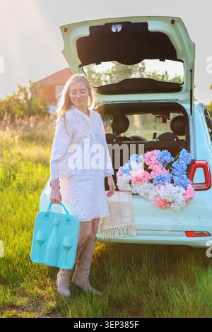 Concetto di stile di vita estivo con una donna sorridente, bagagliaio di auto pieno di fiori e paesaggi di campagna, perfetto per viaggi e svago Foto Stock