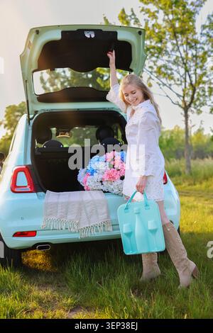 Foto di una donna dallo stile di vita con una custodia turchese in piedi in un prato illuminato dal sole vicino a un'auto piena di fiori, che simboleggia il viaggio e il relax Foto Stock