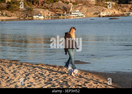 Giovane donna che cammina un piccolo cane bianco lungo la costa sabbiosa di Åros a Søgne, Kristiansand, Norvegia Foto Stock