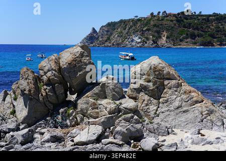 Grandi scogli sulla spiaggia con mare turchese e barche a Santa Maria di Ricadi in Calabria sotto il cielo luminoso e soleggiato dell'Italia meridionale. Foto Stock