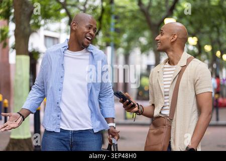Amici maschi africani che camminano e parlano lungo la strada alberata con valigia e smartphone Foto Stock