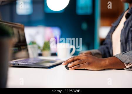 Primo piano di un afroamericano che utilizza un puntatore mentre si lavora sul laptop al tavolo. Messa a fuoco selettiva di una donna nera con un mouse e un computer portatile, che lavora da casa. Foto Stock