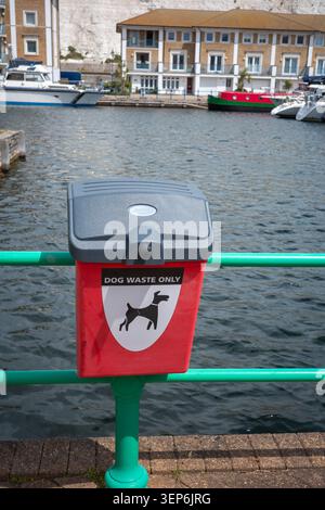 Un contenitore per rifiuti rossi per cani è posizionato vicino all'acqua in un porto. Il contenitore è etichettato solo per i rifiuti di cane. Barche ed edifici possono essere visti dall'altra parte dell'acqua. Foto Stock