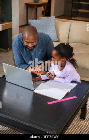Padre e figlio afroamericani che si inclinano in avanti aiutando con la matita e il notebook a casa Foto Stock