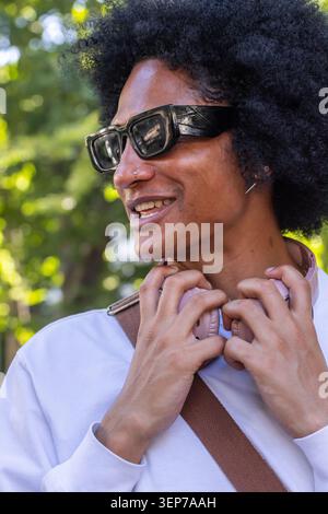 Uomo che tiene le cuffie rosa e sorride indossa tonalità nere, cinturino marrone, anello per il naso nel parco Foto Stock