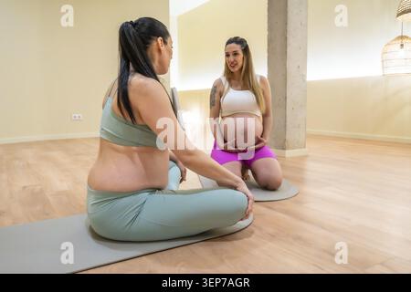 Due donne incinte sedute su tappetini da yoga in uno studio luminoso, sorridenti e chiacchierate mentre condividono la loro routine prenatale di benessere e si sostengono a vicenda Foto Stock