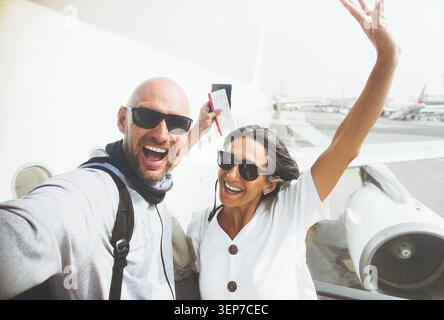 Allegra coppia caucasica eccitata che si mette in posa per selfie con la carta d'imbarco, passaporto davanti all'aereo passeggeri prima di salire a bordo del volo che simboleggia tr Foto Stock