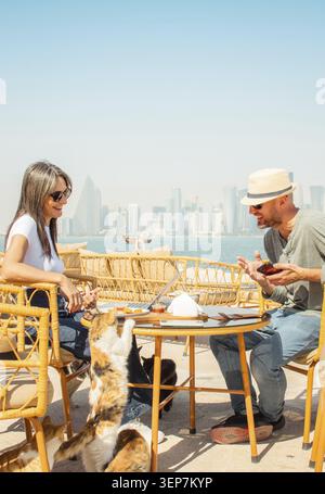Uomo e donna da vicino seduti nel caffè all'aperto Doha Corniche. Donna che gioca a dare da mangiare ai gatti, uomo che usa lo smartphone che si diverte. Sedie Wicker, tavolo, cibo Foto Stock