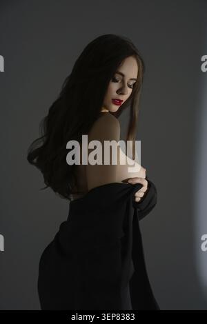 Una donna si trova in uno studio scarsamente illuminato, delicatamente illuminato da una luce delicata. Posa graziosamente con una mano sui suoi vestiti, mettendo in risalto la sua natura Foto Stock