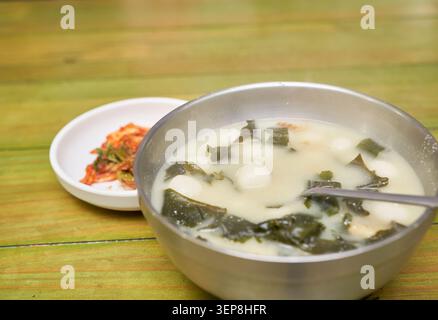 Impasto di riso glutinoso coreano regionale e zuppa di alghe in una ciotola di metallo con un cucchiaio che solleva una palla di riso su un tavolo di legno Foto Stock