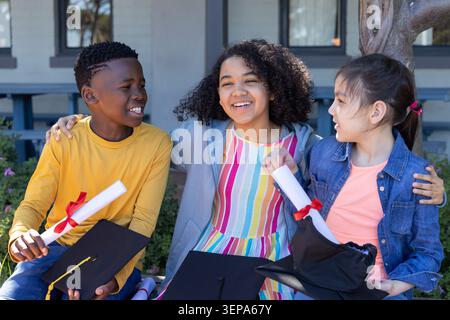 Bambini diversi in abiti luminosi seduti sulla panchina di legno del campus con diplomi a nastro rosso e cappelli Foto Stock