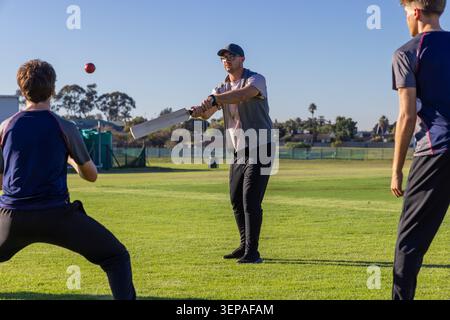 Compagni di squadra maschi che battono e schierano su un campo da cricket erboso in abbigliamento sportivo con mazza, palla rossa, reti Foto Stock