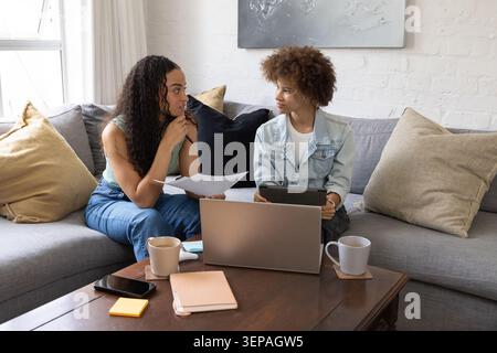 Due donne afroamericane sedute sul divano in soggiorno a discutere di laptop, tablet, appunti stampati Foto Stock