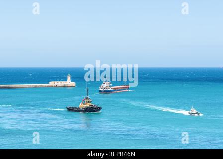 La Valletta, Malta - 16 maggio 2025: Nave da carico, rimorchiatore e pilota navigando vicino all'ingresso storico del porto e al faro di la Valletta, Malta, surro Foto Stock