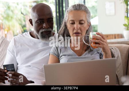 Coppia matura seduta sul divano di casa con laptop, donna con tazza da tè, uomo con carta di credito Foto Stock