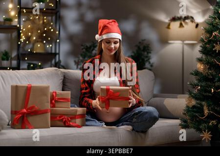 Una donna incinta che indossa un cappello di Babbo Natale apre un regalo di Natale con gioia e anticipazione Foto Stock