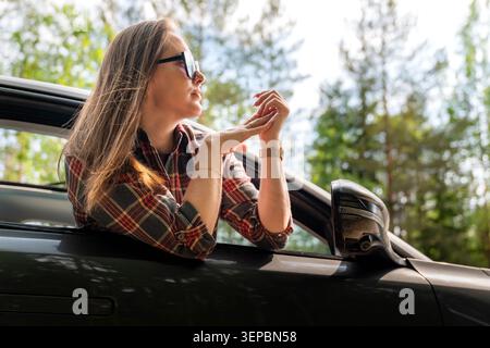 Una giovane donna che indossa occhiali da sole e una camicia a quadri si sporge dal finestrino dell'auto con le mani strette, circondata da alberi verdi e dalla luce naturale brillante Foto Stock