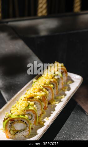 Gli involtini di sushi sono disposti ordinatamente su un lungo piatto in un ristorante. Gli involtini sono riempiti con vari ingredienti e ricoperti di pezzi croccanti. L'atmosfera e' informale e invitante. Foto Stock