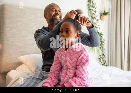 Padre e bambino afroamericani seduti sul letto, padre che usa una spazzola per acconciare i capelli della figlia Foto Stock
