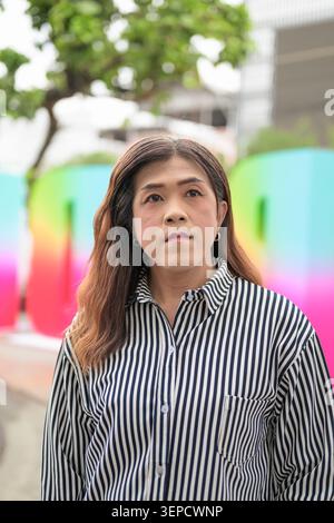 Un ritratto verticale in vita di una donna transgender asiatica di mezza età che guarda lontano mentre posa all'aperto di fronte a un Prid colorato e sfocato Foto Stock