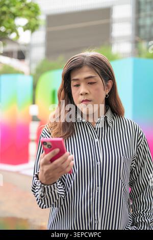 Ritratto verticale in vita di una donna asiatica transgender di mezza età che guarda il suo smartphone all'aperto con uno sfondo sfocato Pride. Foto Stock
