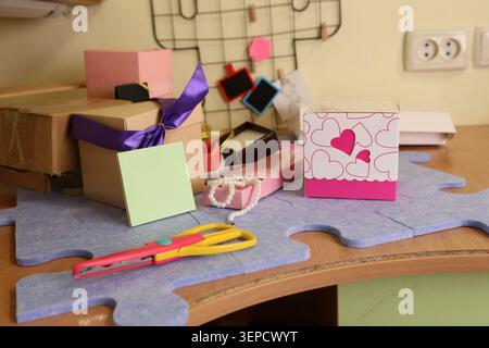 Scatole colorate e carta sono disposte su un tavolo in un piccolo spazio. Le forbici, le note adesive e altri oggetti vengono utilizzati per vari progetti di artigianato. La scena mostra un'area di lavoro creativa. Foto Stock