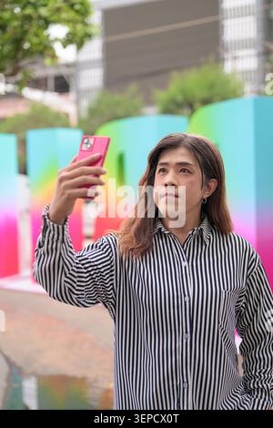 Un ritratto verticale in vita di una donna transgender asiatica di mezza età che scatta un selfie o vlogging all'aperto con uno smartphone rosa contro un colorato Foto Stock