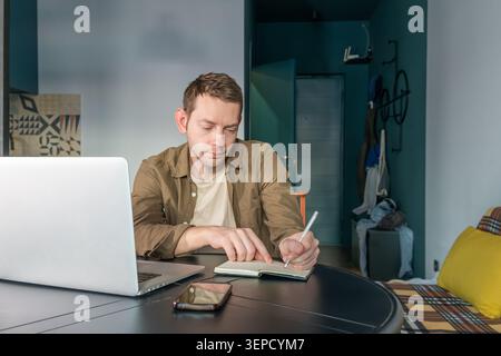 Uomo che scrive appunti su un notebook di carta mentre lavora su un computer portatile a casa. Nomade digitale maschile su lavoro remoto, pianificazione e produttività a casa Foto Stock