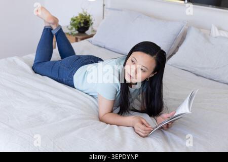 Donna asiatica sdraiata sul letto che legge un libro cartaceo, indossa la parte superiore blu pallido e jeans vicino al comodino Foto Stock