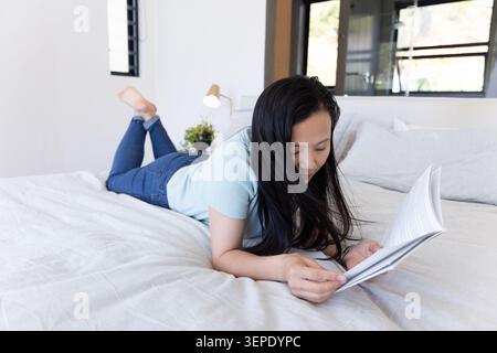 Donna africana matura sdraiata sullo stomaco che legge il libro cartaceo sul letto in una camera da letto luminosa, lampada visibile Foto Stock